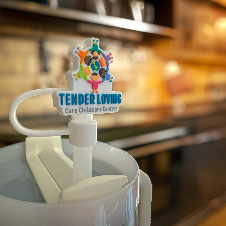 Custom rubber straw topper featuring the Tender Loving Care Childcare Centers logo, made from soft 3D Rubber Bulk PVC material, attached to a straw in a drink container, with a blurred café background.