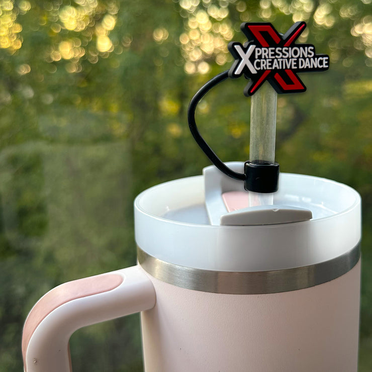 Custom rubber straw topper featuring the Xpressions Creative Dance logo in red, black, and white, made from flexible PVC material, attached to a straw in a pink Stanley tumbler with a scenic outdoor background.