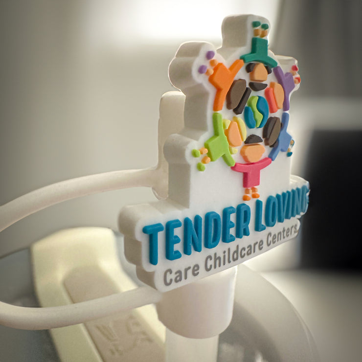 Close-up of a custom rubber straw topper featuring the Tender Loving Care Childcare Centers logo, made from soft PVC material, attached to a straw in a Stanley Tumbler with a blurred indoor background.