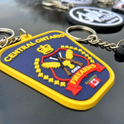 Custom PVC promotional keychain featuring the Central Ontario Firearms Academy logo, with raised 3D design, yellow border, and embossed text, attached to a metal keyring on a reflective surface.